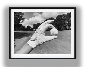 Op de afbeelding: Een zwart-witfoto van een golfhandschoen die een golfbal met putjes vasthoudt. De handschoen is licht van kleur met zichtbare stiksels en een polssluiting, geplaatst tegen een wazige achtergrond van een golfbaan met bomen en wolken.