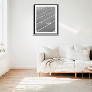 May include: A black and white framed wall art print featuring a high-angle view of a running track with white lane markings. The image shows the curved lines of the track surface extending across the frame, captured in a monochromatic style.