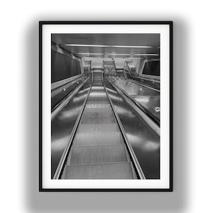 May include: Black and white photograph of a framed escalator. The escalator is in a public space, with metal steps and handrails. The image is a high-angle shot, looking down the length of the escalator.