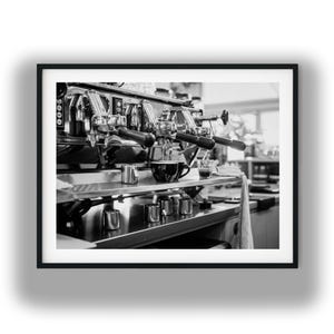 Puede incluir: Una fotografía en blanco y negro de una máquina de café expreso profesional con varios grupos y portafiltros. Una taza de cerámica oscura descansa sobre la bandeja, mientras varias jarras de metal pequeñas están en la encimera.