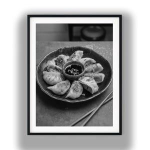 May include: Black and white photograph of a plate of dumplings with a small bowl of dipping sauce in the center. The dumplings are arranged around the bowl, and chopsticks are placed on the side of the plate. The image is framed in a black border.