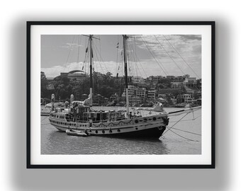May include: Black and white photograph of a sailboat docked in a harbor. The boat has two masts and is surrounded by water. Buildings and trees are visible in the background, under a cloudy sky.