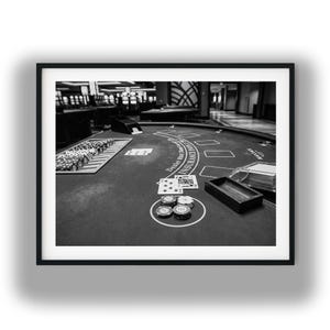 May include: A monochrome image of a blackjack table in a casino setting. The table features playing cards, casino chips, and a wooden chip tray. The text "Dealer Must Stand" is visible on the green felt.