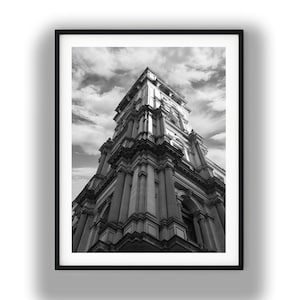 May include: Black and white framed print of a tall building with ornate architectural details. The building has multiple levels, columns, and arched windows. The sky is filled with clouds, creating a dramatic contrast.