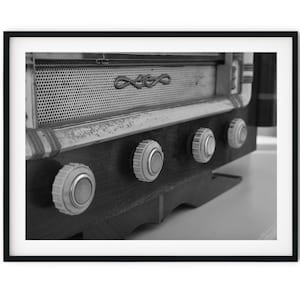 Puede incluir: Una radio vintage en blanco y negro con tres botones blancos. La radio tiene una rejilla metálica y una base de madera.