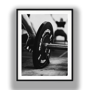 Pode incluir: Uma fotografia a preto e branco com uma barra de pesos e um disco preto com a inscrição REP FITNESS 15LB. O disco está colocado no chão de um ginásio, com uma mão a segurar a barra. No fundo desfocado, uma figura está a levantar pesos.