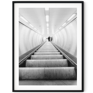 May include: A black and white photograph of a long escalator in a subway station. The escalator is empty except for a single person walking up the stairs.