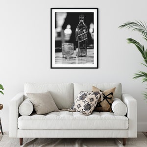 May include: A black and white framed wall art print featuring a bottle of Johnnie Walker Black Label scotch whisky and a glass filled with liquid and ice cubes, displayed in a simple black frame on a plain white wall above a white sofa.