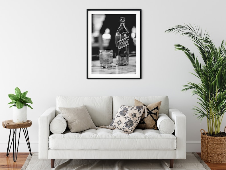 May include: A black and white framed wall art print featuring a bottle of Johnnie Walker Black Label scotch whisky and a glass filled with liquid and ice cubes, displayed in a simple black frame on a plain white wall above a white sofa.
