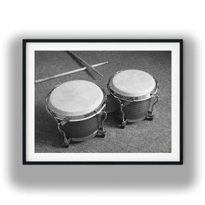 May include: A black and white photograph of a set of bongo drums with two drumsticks resting diagonally across the top. The drums feature metallic rims and light-colored drumheads, presented within a simple black frame for display.
