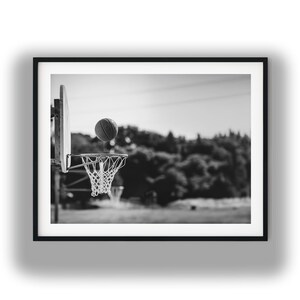 May include: Black and white framed photograph of a basketball in mid-air, about to go through the net. The basketball hoop is mounted on a wooden backboard. The background is blurred, showing trees and a field.