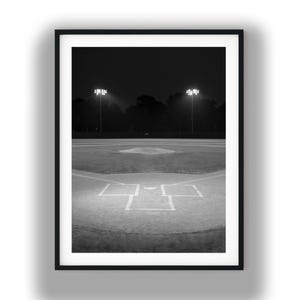May include: A black and white framed photograph of a baseball field at night. The image shows the home plate area with white chalk lines on the dirt, illuminated by two tall stadium light poles standing against a dark, silhouetted tree line.