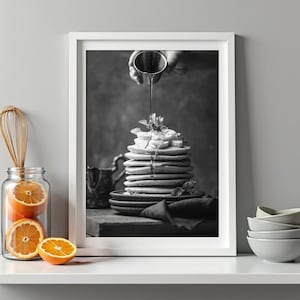 Puede incluir: Una impresión de arte mural en blanco y negro enmarcada que muestra una alta pila de tortitas cubiertas con rodajas de plátano y hojas de menta, con sirope vertiéndose desde una pequeña jarra de metal, sobre una superficie de madera oscura.