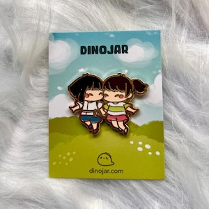 May include: Gold enamel pin featuring two cartoon girls with black hair, one wearing a white and blue outfit and the other wearing a green and white outfit. The pin is on a white card with the text "DINOJAR" and "dinojar.com".