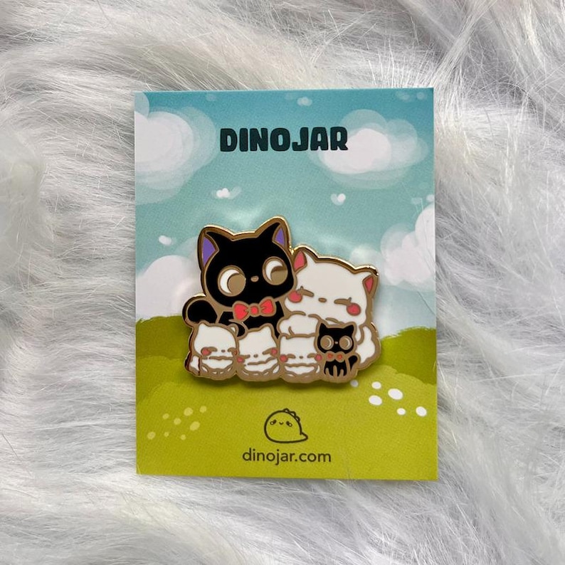 May include: A black cat with a red bow tie and white cats with black eyes are featured on a gold enamel pin. The pin is on a green and white card with the text "Dinojar" and "dinojar.com".