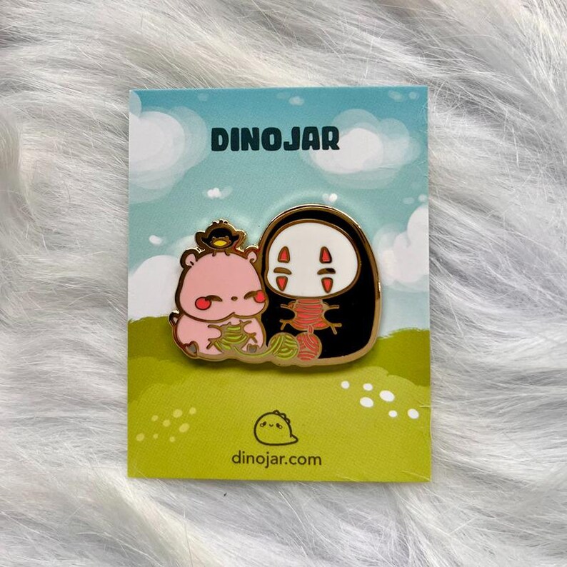 May include: A gold enamel pin featuring a pink cartoon character with a bird on its head and a black No Face character from the movie Spirited Away. Both characters are holding yarn balls. The pin is on a white card with the text "Dinojar" and "dinojar.com".