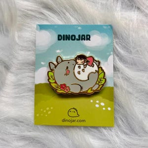 May include: A gold enamel pin featuring a cartoon character with a pink dress sleeping on a gray and white fluffy creature. The pin is surrounded by green leaves and pink flowers. The pin is on a white card with the text "DINOJAR" and "dinojar.com".
