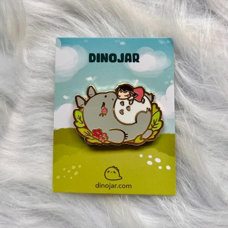 May include: A gold enamel pin featuring a cartoon character with a pink dress sleeping on a gray and white fluffy creature. The pin is surrounded by green leaves and pink flowers. The pin is on a white card with the text "DINOJAR" and "dinojar.com".
