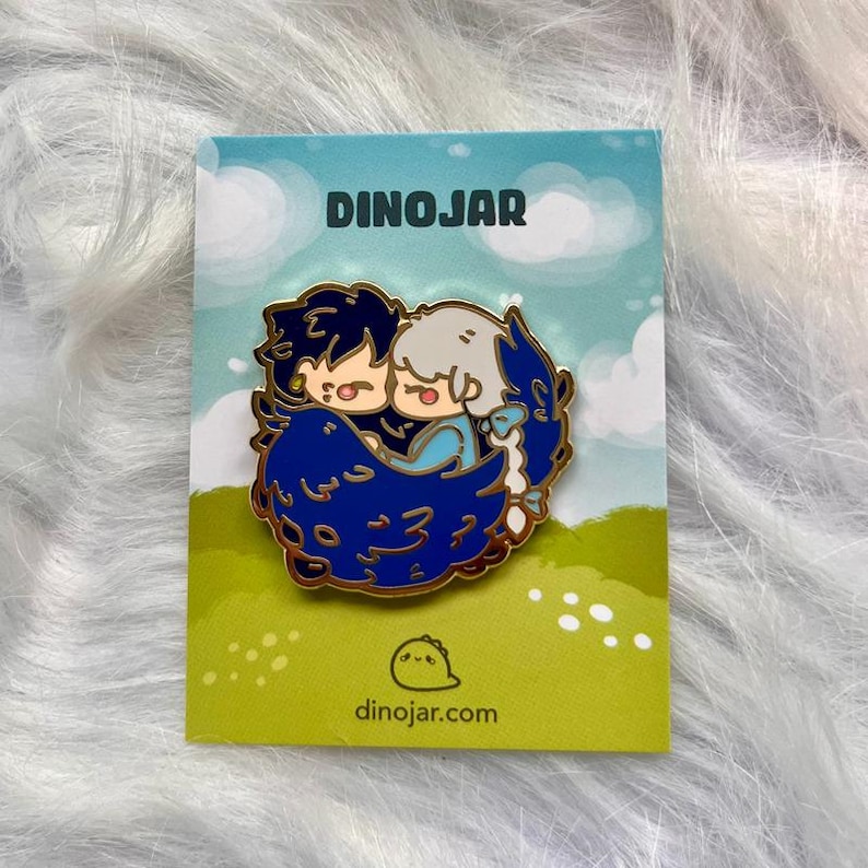 May include: A gold enamel pin featuring two cartoon characters, one with blue hair and the other with white hair, hugging each other. The characters are surrounded by a blue and gold design. The pin is on a white card with the text "DINOJAR" and "dinojar.com".