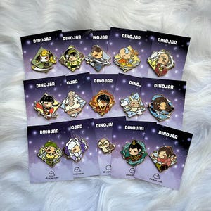 May include: A set of 16 enamel pins featuring characters from the animated television series Avatar: The Last Airbender. The pins are displayed on individual cards with a dark blue background and white stars. The characters include Aang, Katara, Sokka, Toph, Zuko, and Azula.