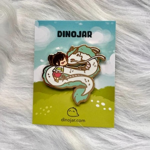 May include: Gold enamel pin featuring a cartoon girl hugging a white and green dragon. The pin has a white background with green grass and white dots. The text "DINOJAR" is at the top of the pin and "dinojar.com" is at the bottom.