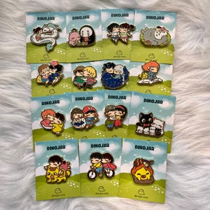 May include: A collection of 12 enamel pins featuring characters from popular animated films. The pins are colorful and feature a variety of characters, including Totoro, Kiki's Delivery Service, and Spirited Away. The pins are all set against a green background with the word "DINOJAR" printed on each card.