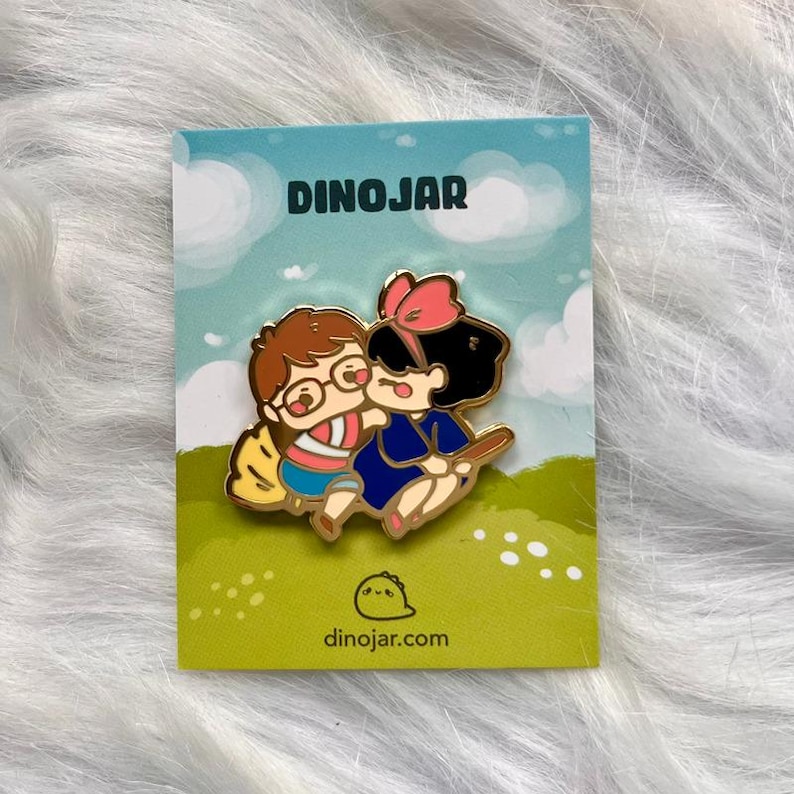 May include: A gold enamel pin featuring a cartoon illustration of two characters, a girl with black hair and a red bow, and a boy with brown hair and glasses, riding a broomstick. The pin is on a white card with the text "DINOJAR" and "dinojar.com".