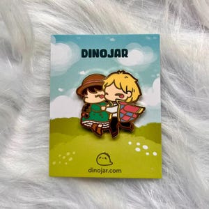 May include: A gold-colored enamel pin featuring a cartoon couple hugging. The woman is wearing a green dress and a brown hat. The man is wearing a blue and red patterned cloak. The pin is on a white card with the text "Dinojar" and "dinojar.com".