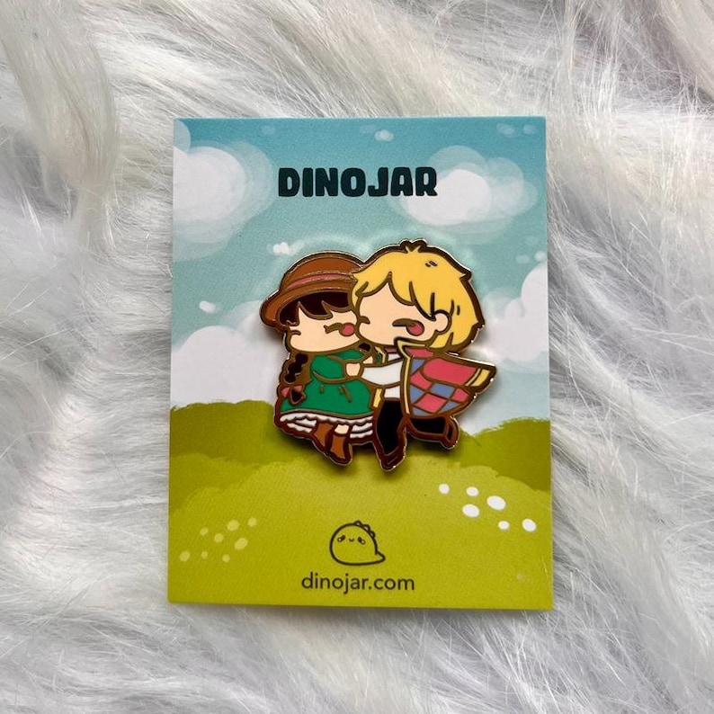 May include: A gold-colored enamel pin featuring a cartoon couple hugging. The woman is wearing a green dress and a brown hat. The man is wearing a blue and red patterned cloak. The pin is on a white card with the text "Dinojar" and "dinojar.com".