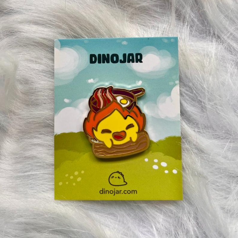 May include: A gold enamel pin featuring a cartoon dinosaur with a yellow body, orange flames, and a red and white bacon and egg breakfast on its head. The pin is on a white card with the text "Dinojar" and "dinojar.com".