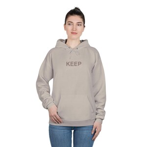 May include: A beige hoodie with the words "KEEP going" printed on the front. The hoodie features a drawstring hood and a front pocket. The person is wearing blue jeans.