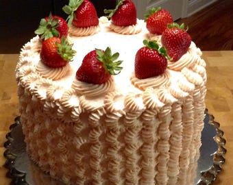 Puede incluir: Un pastel redondo con glaseado de color melocotón claro, decorado con volantes verticales en los laterales y remolinos en la parte superior. Siete fresas rojas frescas enteras con tallos verdes están dispuestas en círculo sobre el pastel.