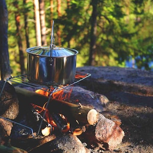May include: A stainless steel pot with a handle sits on a metal grate over a campfire. The pot is sitting on a bed of logs and embers.