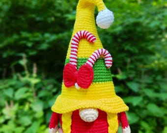 May include: A crocheted gnome figure featuring a tall yellow hat with a white pom-pom, a green band, and two red and white striped candy cane accents with a red bow. The gnome has a round white nose, yellow braided hair, and a red body.