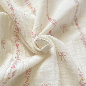 May include: Cream-coloured fabric with a floral and bow design in pink, yellow, and green. The fabric is soft and draped, with a subtle texture. The design features repeating vertical borders of flowers and bows.