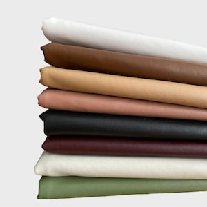 May include: A stack of folded fabric in various colors, including white, brown, tan, pink, black, burgundy, off-white, and green. The fabric appears to be leather or a leather-like material, with a smooth texture and a slight sheen.