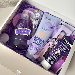 Könnte beinhalten: Eine weiße Geschenkbox gefüllt mit lila Papierstreifen, die eine lila Pumpspenderflasche mit einem Good Things Take Time Aufkleber, eine Jean & Len Mango Maracuja Duschgel-Tube, eine Lavera Handcreme-Tube, eine runde Balea-Dose und einen Schlüsselanhänger enthält.