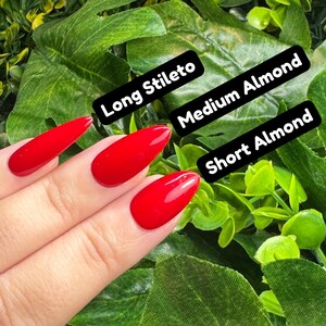 K&ouml;nnte beinhalten: Eine Hand zeigt drei rote Nagelformen: Long Stiletto, Medium Almond und Short Almond. Die N&auml;gel sind gl&auml;nzend rot lackiert. Der Hintergrund zeigt gr&uuml;nes Laub.