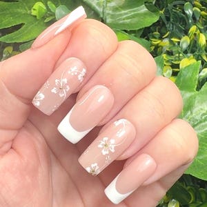 K&ouml;nnte beinhalten: Eine Hand, die lange, quadratisch geformte N&auml;gel zeigt. Einige N&auml;gel haben ein French-Manik&uuml;re-Design mit wei&szlig;en Spitzen, w&auml;hrend andere eine nackte Basis mit zarten wei&szlig;en Blumenmustern aufweisen. Die N&auml;gel sind gut gepflegt und poliert.