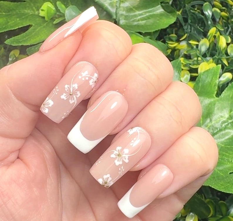 K&ouml;nnte beinhalten: Eine Hand, die lange, quadratisch geformte N&auml;gel zeigt. Einige N&auml;gel haben ein French-Manik&uuml;re-Design mit wei&szlig;en Spitzen, w&auml;hrend andere eine nackte Basis mit zarten wei&szlig;en Blumenmustern aufweisen. Die N&auml;gel sind gut gepflegt und poliert.