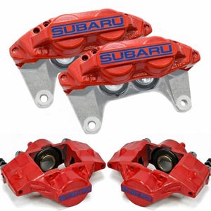 May include: Four red automotive brake calipers featuring the blue Subaru logo. The top two calipers show the mounting points and silver metal finish, while the bottom two show the internal piston housing and the Subaru text on the front face.