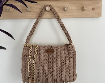 May include: A brown crocheted handbag with a gold chain strap. The bag has a small brown leather tag with the text "Knit Addict" on it.