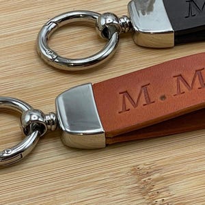 Könnte beinhalten: Zwei Leder-Schlüsselanhänger mit silberfarbener Hardware. Einer ist braun mit geprägtem "M.M", der andere ist schwarz mit geprägtem "M". Die Schlüsselanhänger haben einen Metallverschluss und ein rechteckiges Lederetikett.