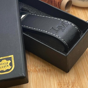 Könnte beinhalten: Ein schwarzer Leder-Geldscheinclip mit weißen Nähten und den Initialen "LM" auf der Vorderseite, präsentiert in einer schwarzen Geschenkbox. Die Box hat ein goldenes Emblem mit der Aufschrift "ECHTES LEDER".