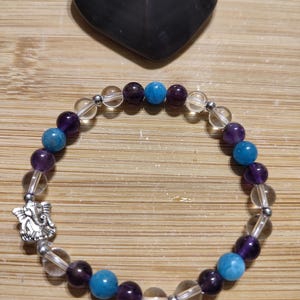May include: A bracelet made with clear, amethyst, and blue beads. The bracelet has a silver charm in the shape of an angel.