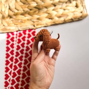 May include: A small brown crocheted dachshund dog keychain. The keychain is being held in a hand with a red and white bracelet.