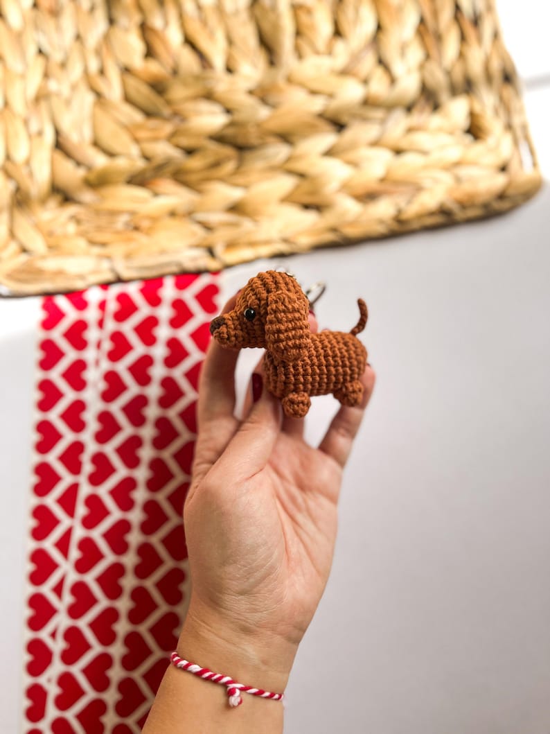 May include: A small brown crocheted dachshund dog keychain. The keychain is being held in a hand with a red and white bracelet.