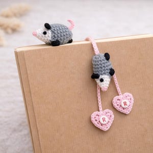 May include: A handmade crochet bookmark with a grey and white opossum design. The bookmark has a pink cord with two pink heart-shaped charms, each with a letter. The opossum is on a brown book.