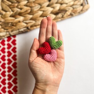 May include: Three small crocheted hearts in different colors: red, green, and pink. The hearts are held in a person's hand.