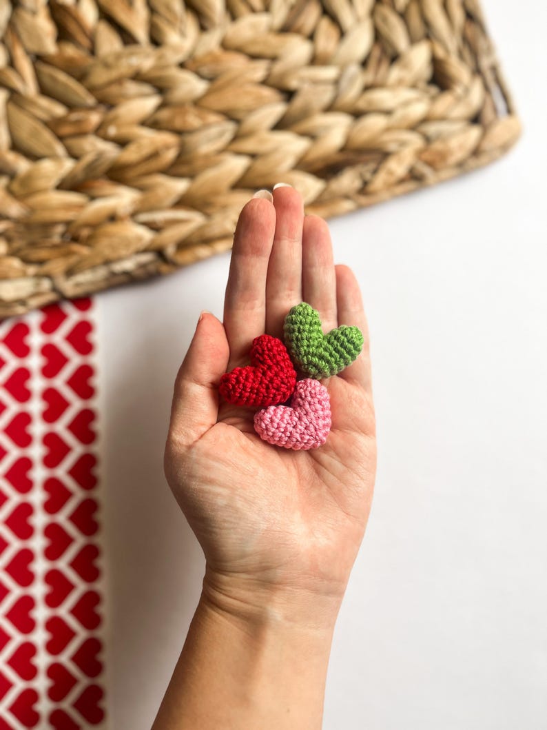 May include: Three small crocheted hearts in different colors: red, green, and pink. The hearts are held in a person's hand.